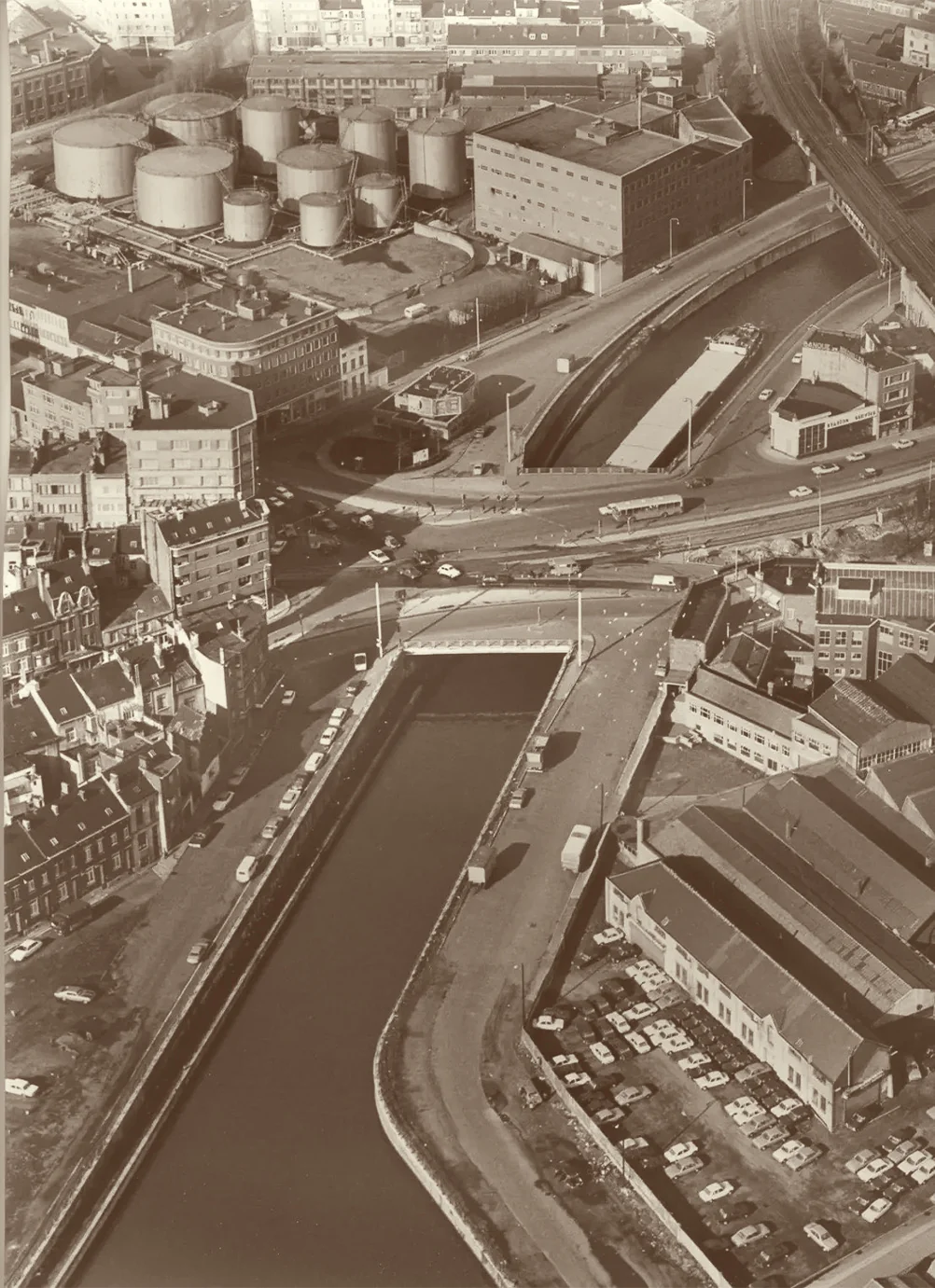 Photo aérienne des années 1960 : les grandes cuves de Shell et leurs entrepôts au bord du canal, un pont et des voitures garées sur les quais
