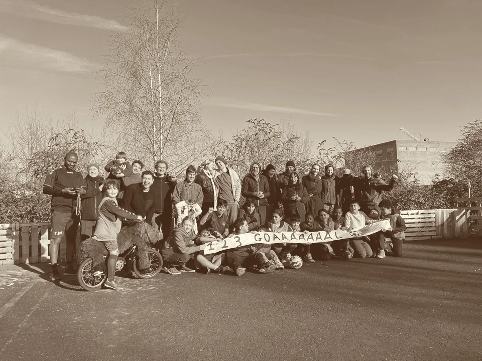 Photo de groupe sur le terrain de foot improvisé : enfants et adultes du quartier réunis derrière une banderole, sur l’ancien parking asphalté bordé de palettes