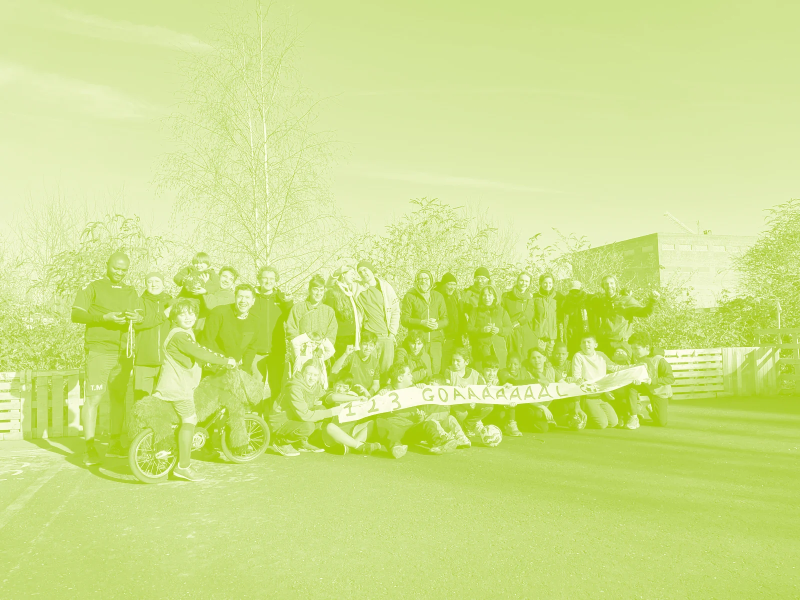 Photo de groupe sur le terrain de foot improvisé : enfants et adultes du quartier réunis derrière une banderole, sur l’ancien parking asphalté bordé de palettes