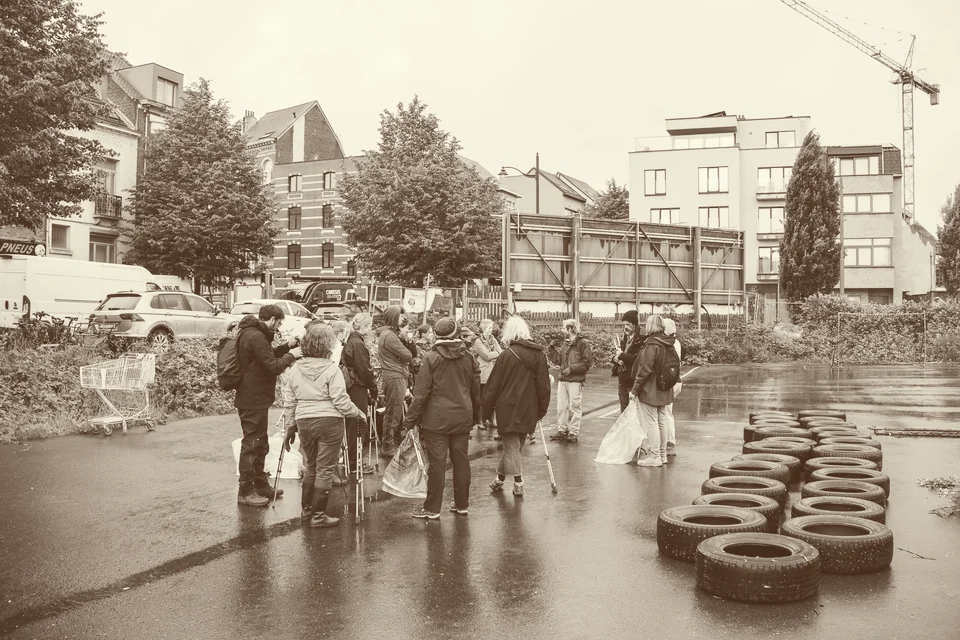 Crade party sur le parking asphalté du marais : un groupe de bénévoles avec des sacs et des pinces à déchets, des pneus empilés, des immeubles et une grue à l’arrière-plan