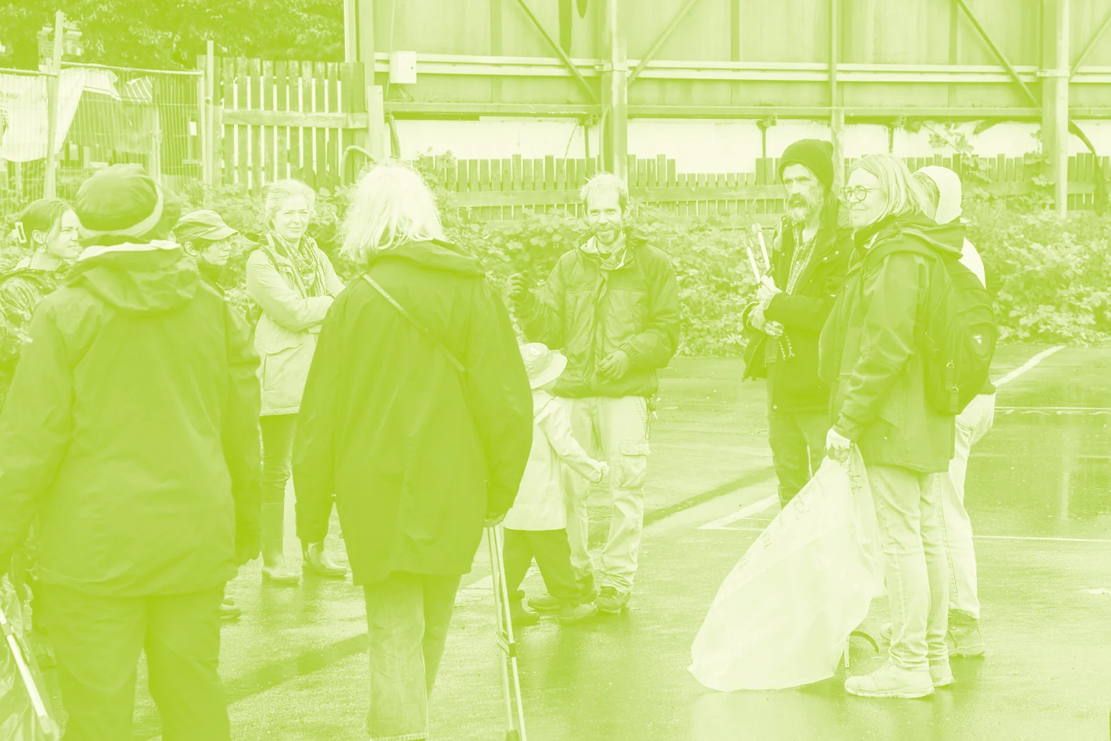 Bénévoles de la crade party en vestes de pluie, pinces et sacs à déchets à la main, qui discutent sur le parking mouillé devant les palissades du site