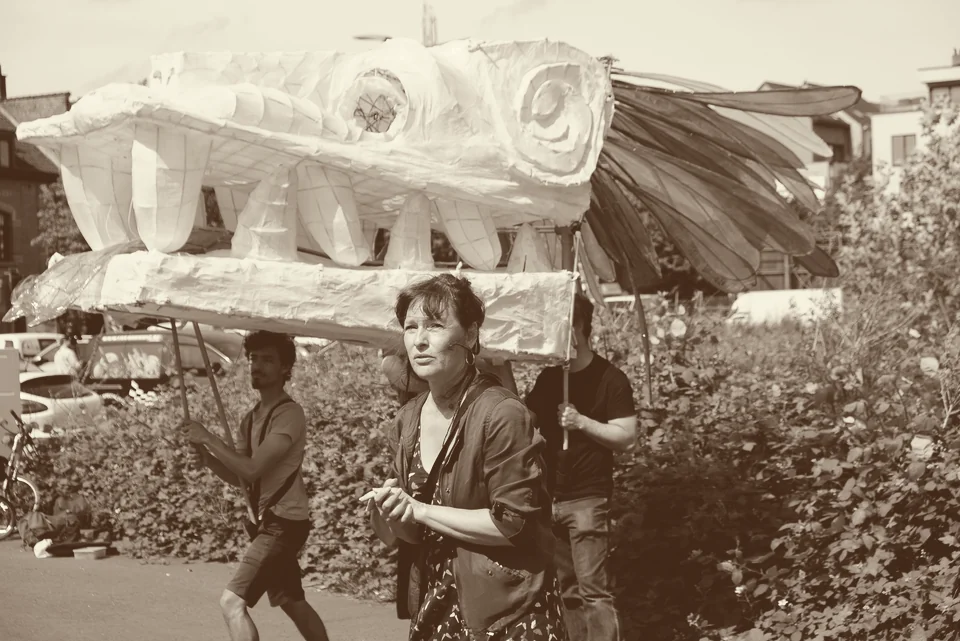 Une grande tête de dragon en papier mâché, aux ailes déployées, portée à bout de perches par plusieurs personnes devant la végétation du marais ; une femme marche au premier plan