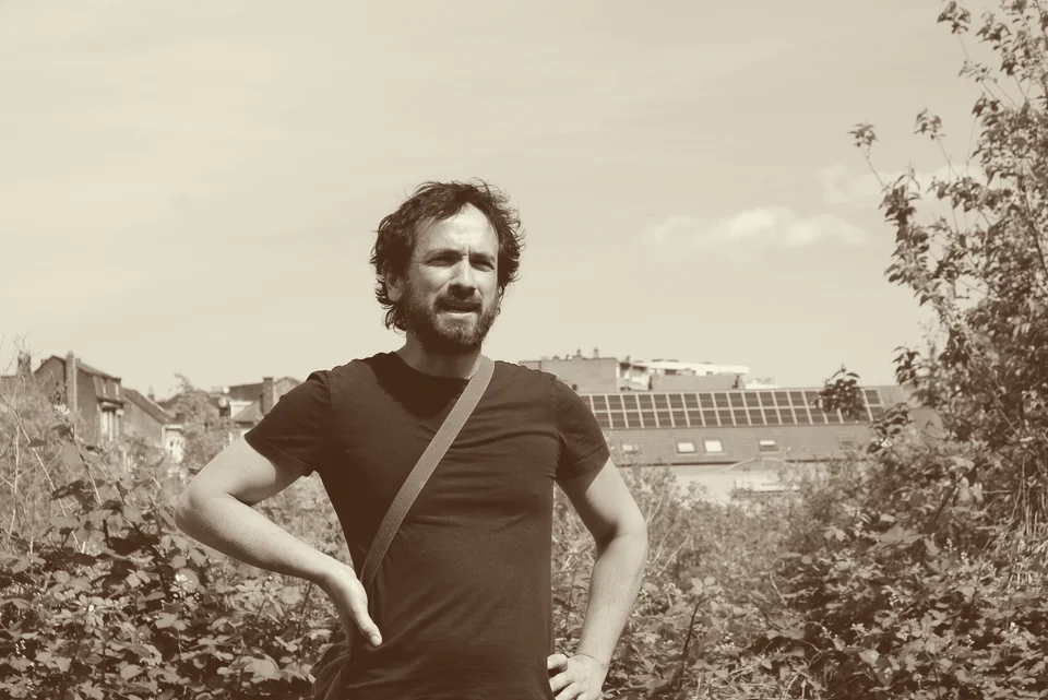 Portrait d’un défenseur du marais, barbu, en tee-shirt, les mains sur les hanches, debout dans la végétation du site, des toits d’Anderlecht à l’arrière-plan