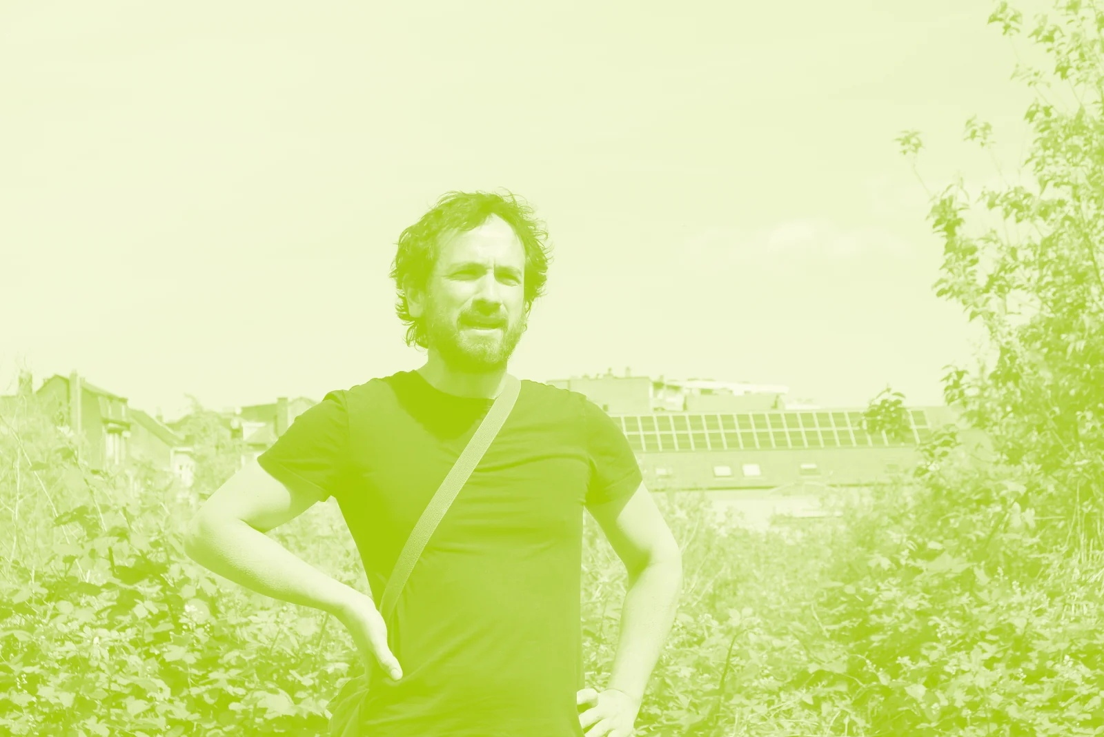 Portrait d’un défenseur du marais, barbu, en tee-shirt, les mains sur les hanches, debout dans la végétation du site, des toits d’Anderlecht à l’arrière-plan
